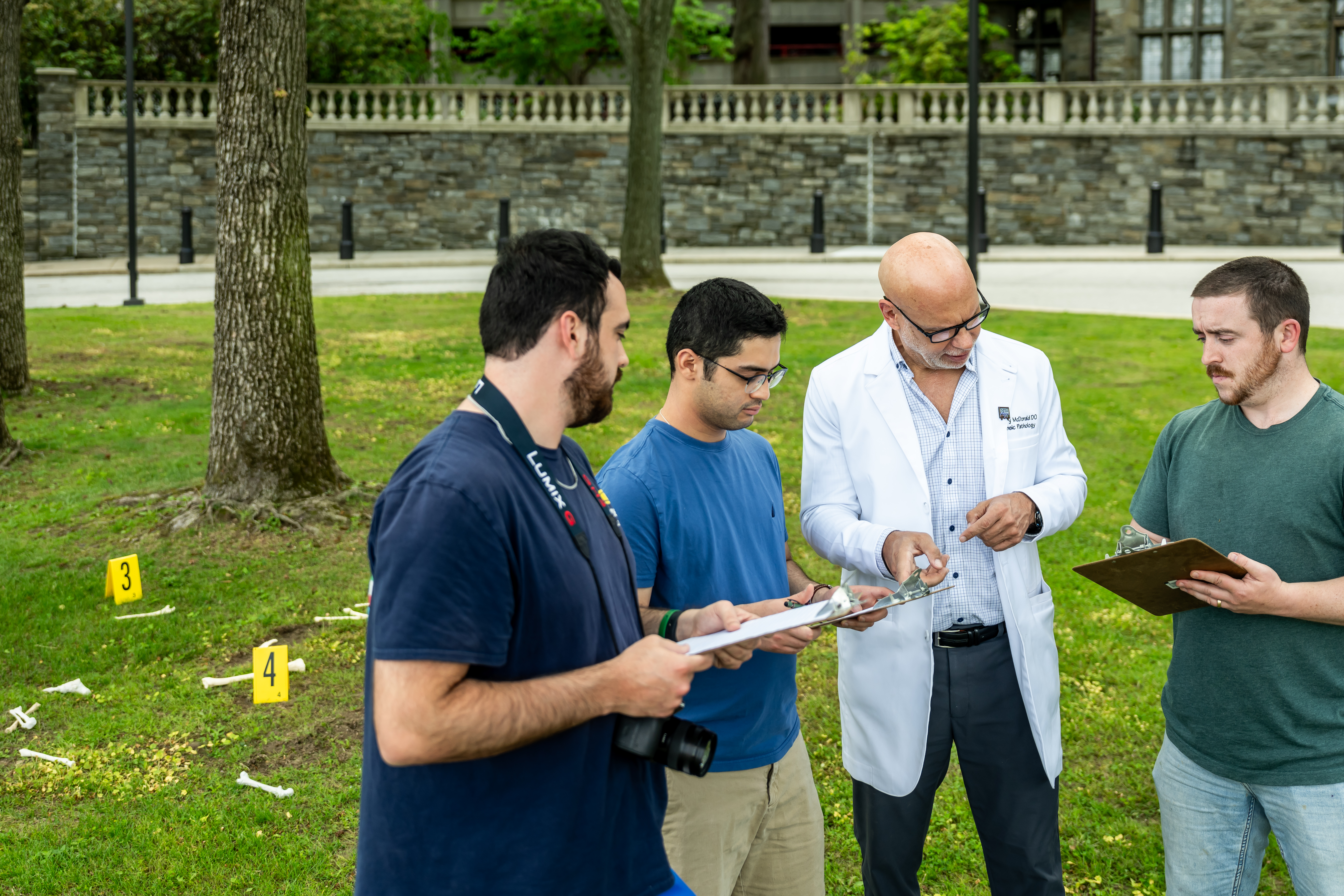forensic medicine students gather with Greg McDonald in a white lab coat to compare notes outside at a crime scene reinactment
