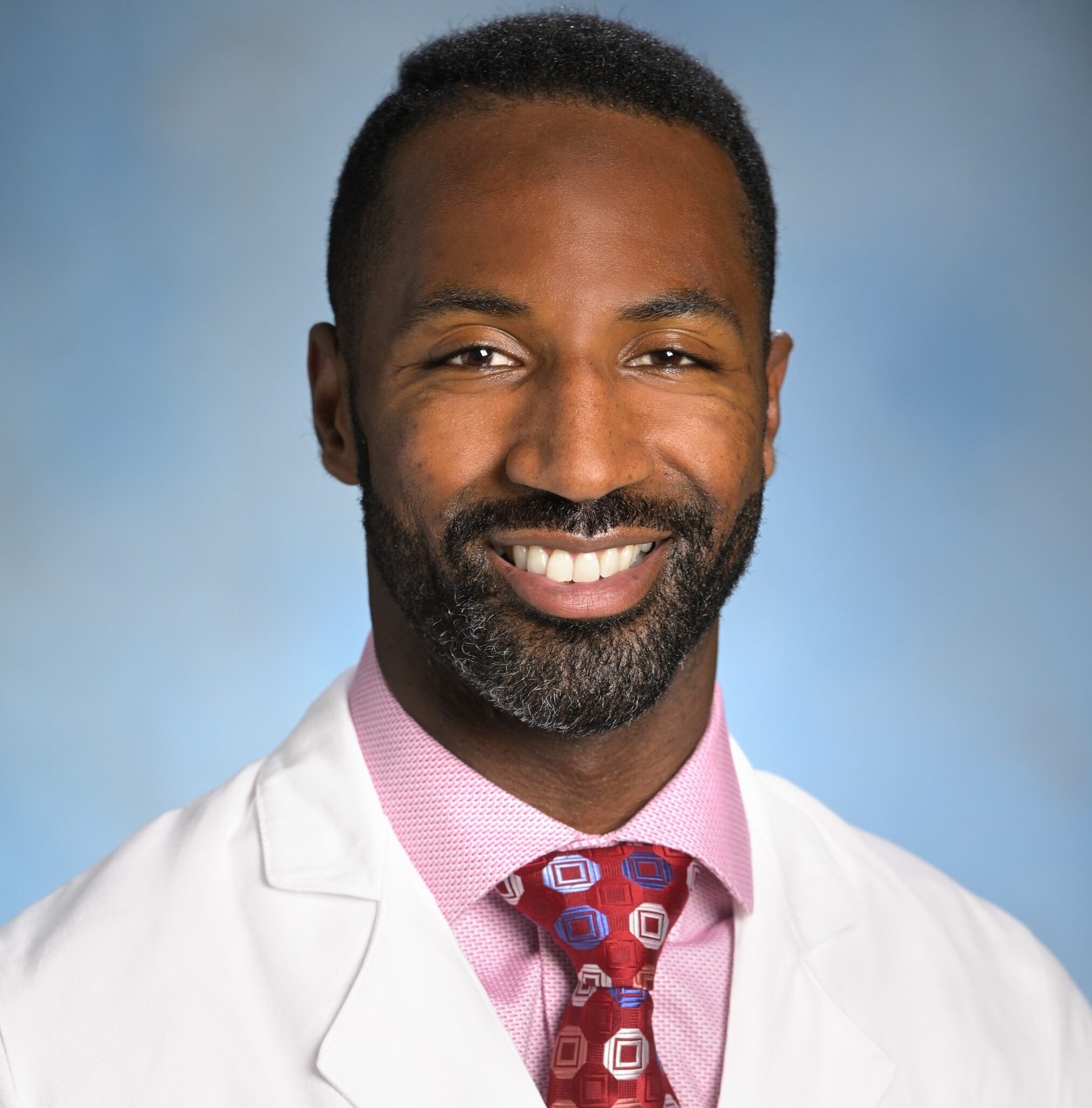 Dominique Brundidge smiles in a pink collared shirt, red tie, and Main Line Health white coat against a light blue background