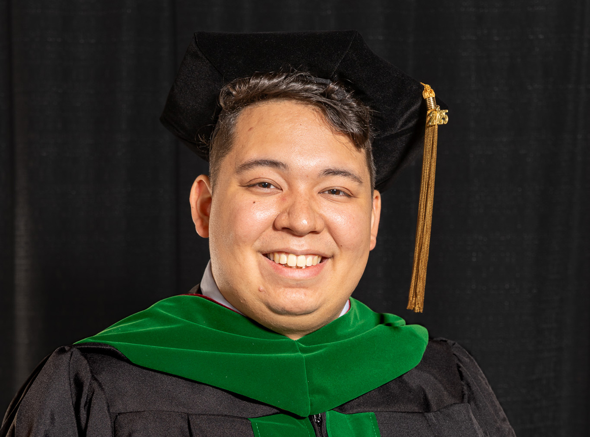 a male with brown hair smiles in commencement regalia against a black background