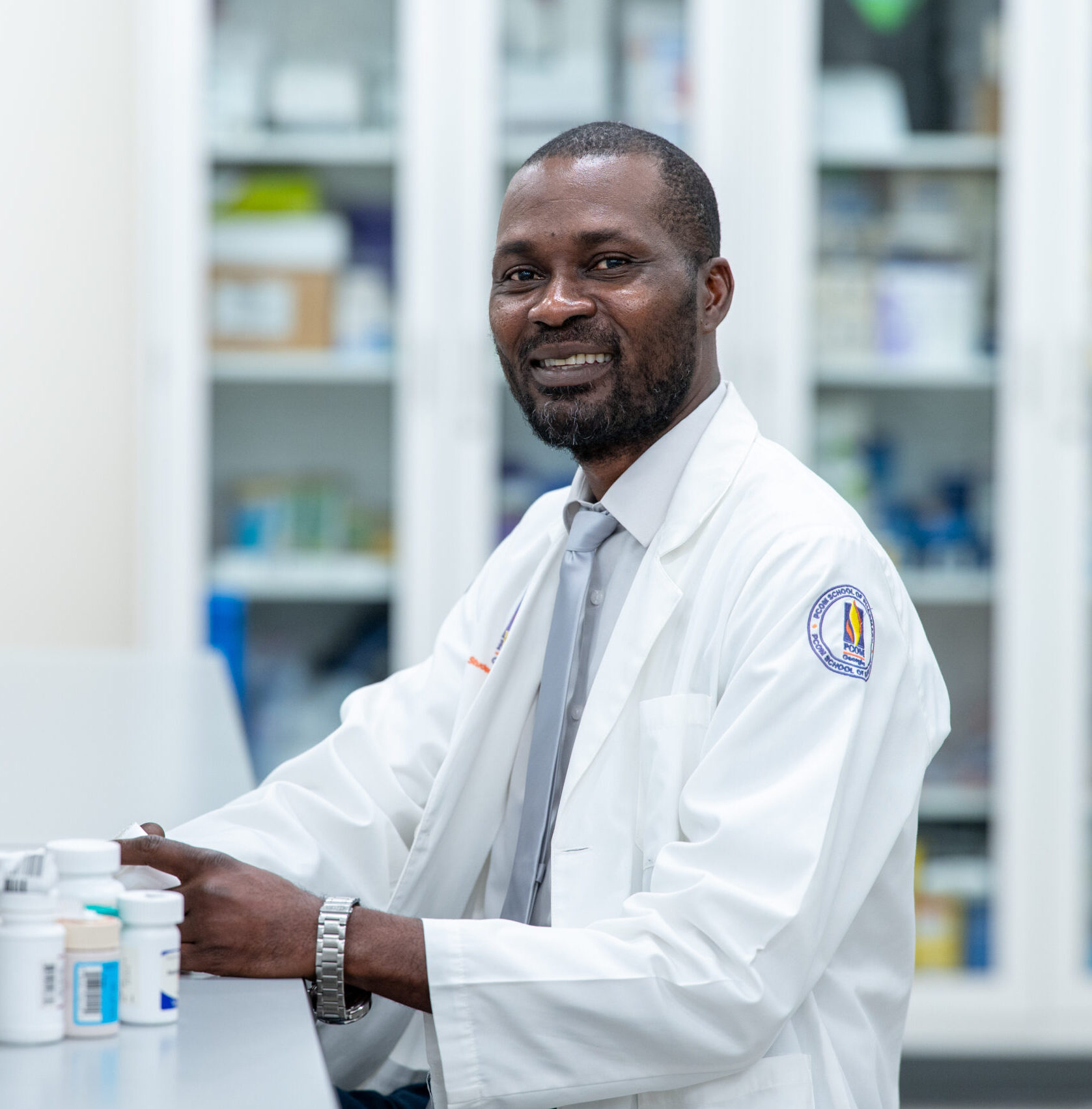 A male pharmacy student smiles wearing a white coat and working with prescription bottles with the pharmacy lab in the background