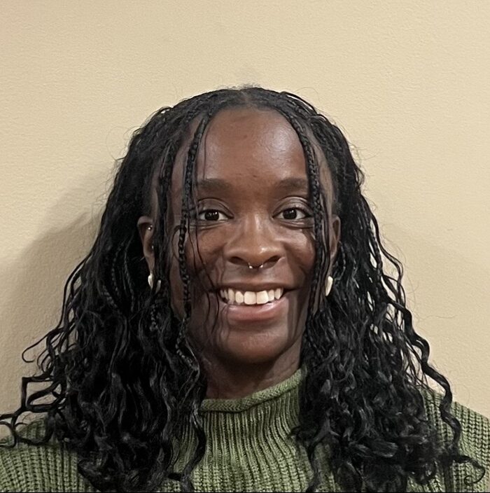 A female student in a green shirt smiles while standing against a beige backgroudn