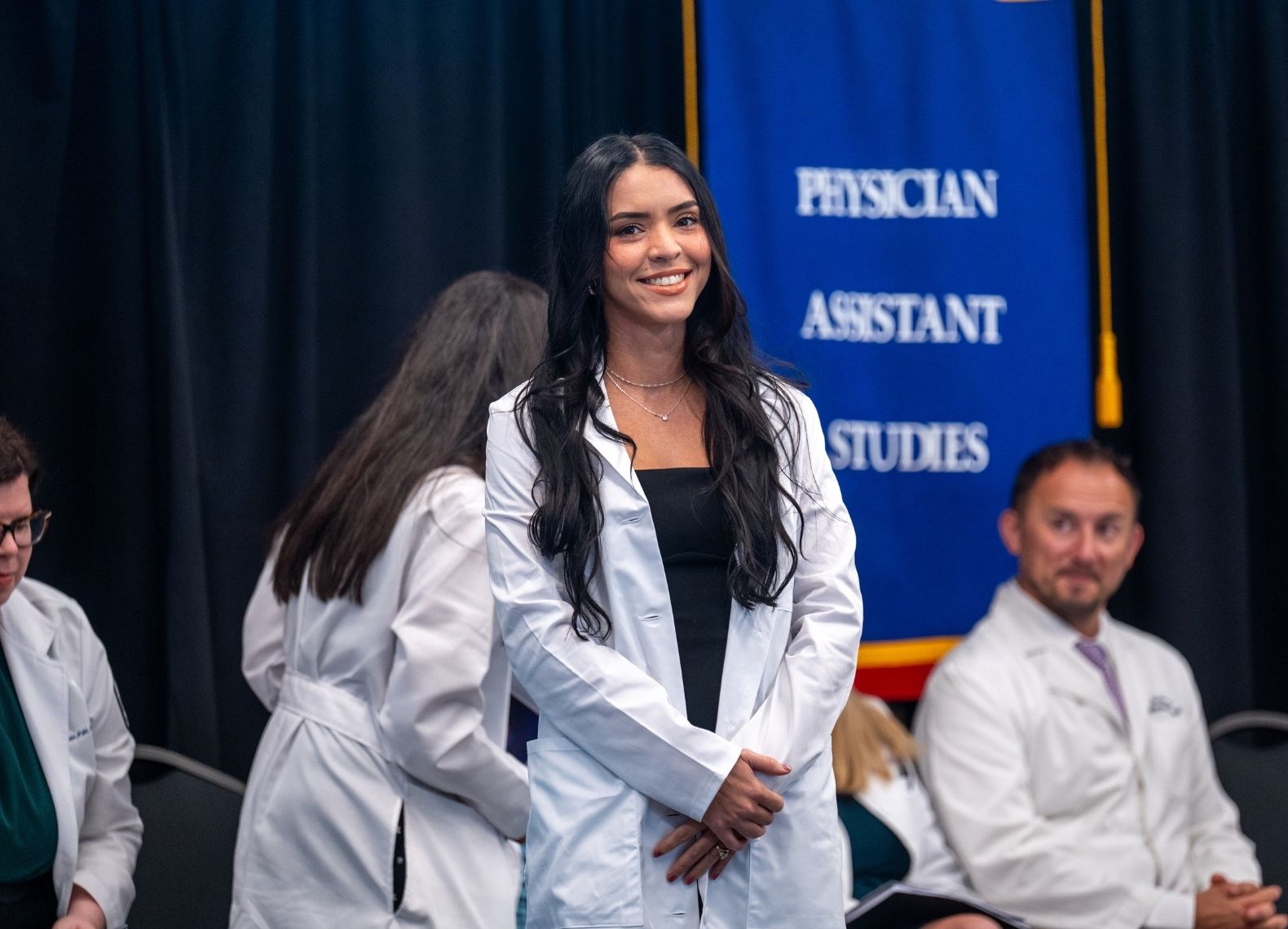 Smiling physician assistant student stands on stage in a new white coat at a PCOM PA Studies white coat ceremony