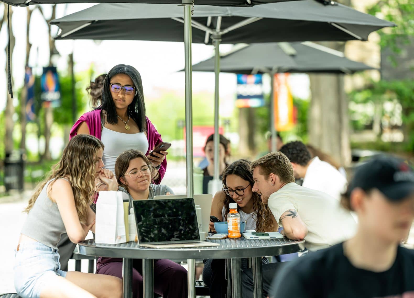 A group of students gather around an outdoor umbrella table with laptops and snacks on the campus plaza