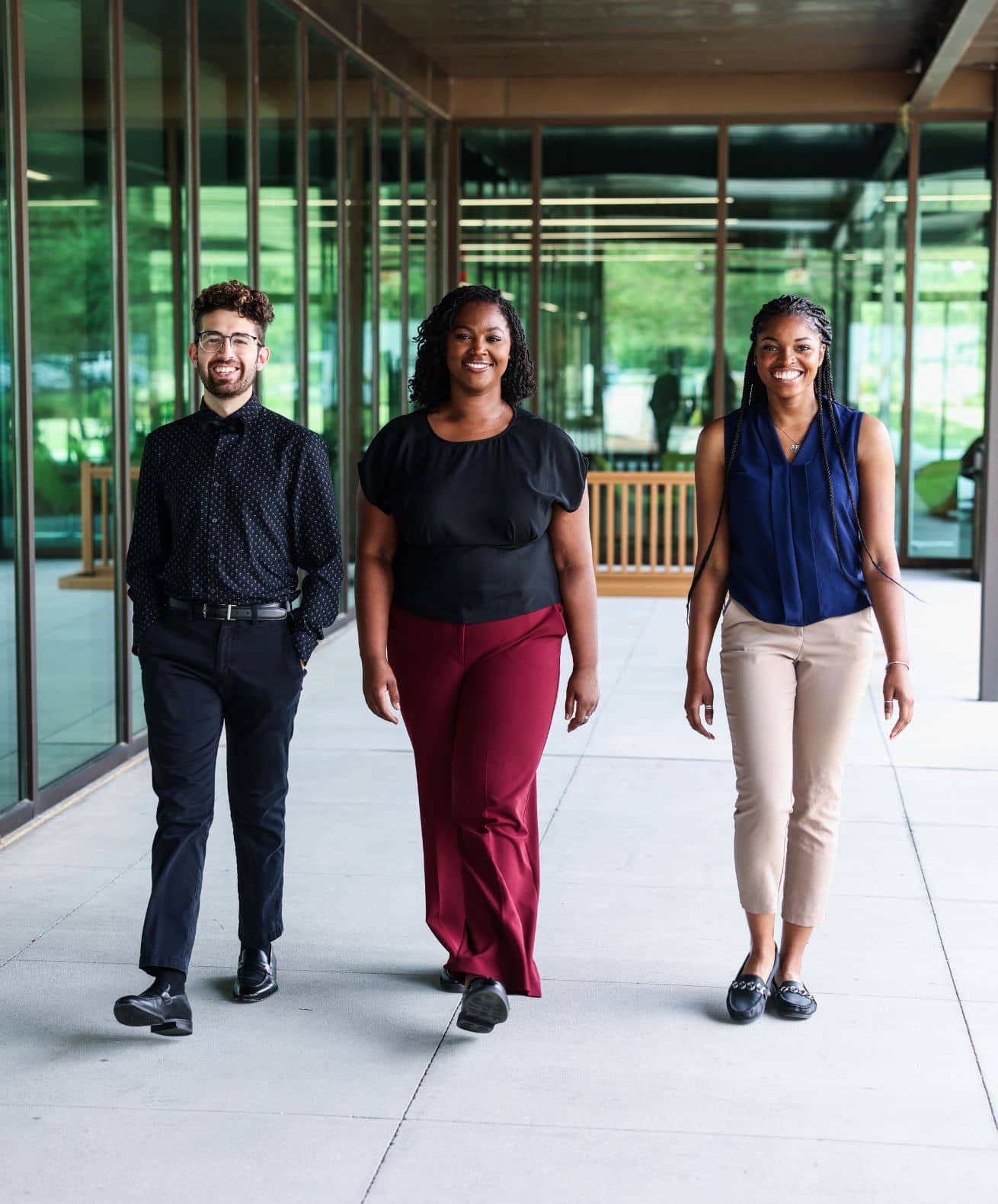 Three smiling professionals walk side by side through a modern glass-walled campus corridor