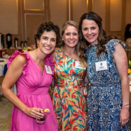 Three women in colorful dresses wearing name tags smile together at a PCOM networking event